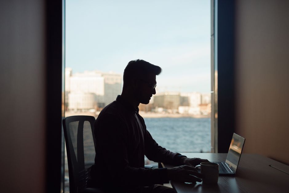 Silhouette of a man working on his laptop by a window overlooking a waterfront cityscape.