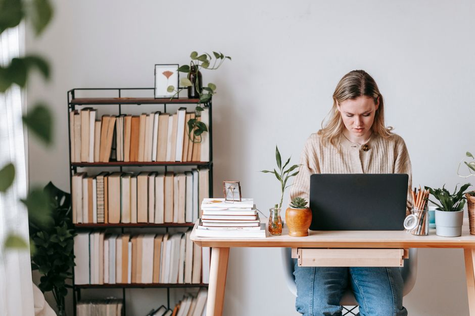 Focused female remote employee working on project on netbook at table with potted plants against bookcase at home on light background