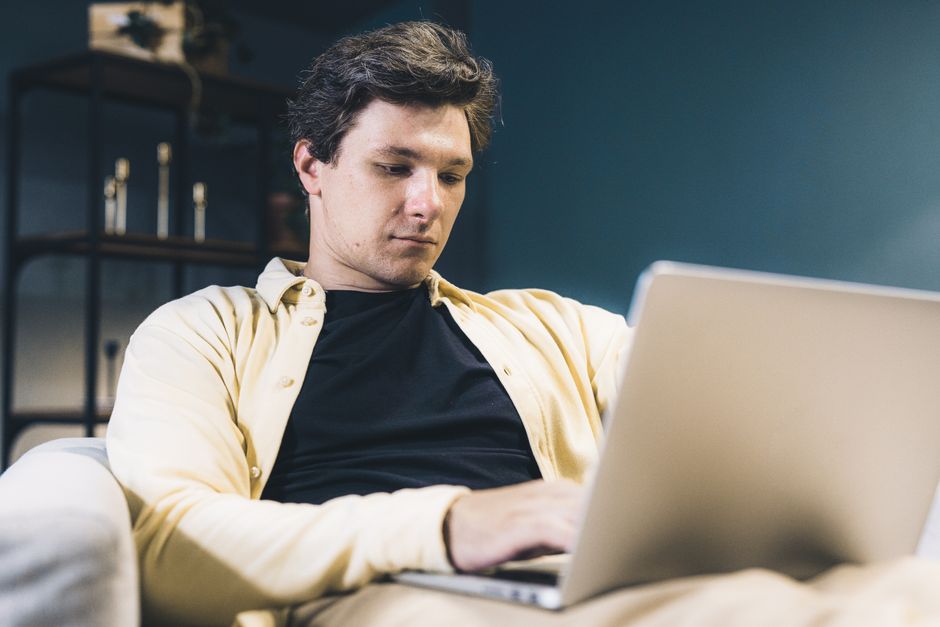Focused young man sitting comfortably using a laptop at home, engaging in remote work.